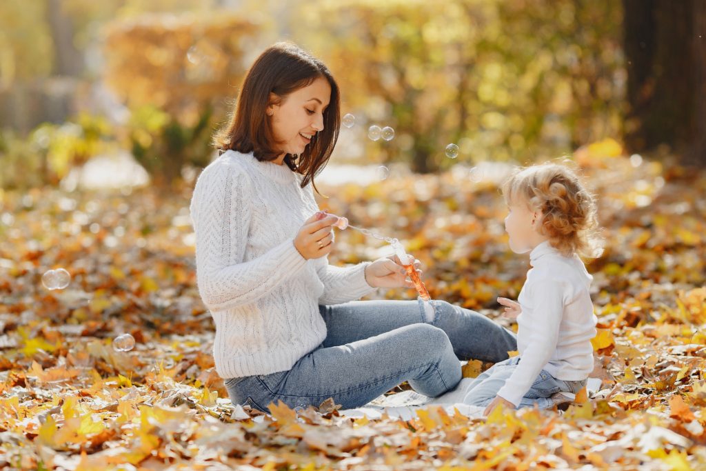 A woman and a young female child are sitting on a leave covered ground with bubbles floating around. Nurturing mindfulness