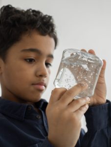 Young male child holding a clear glass container that is filled with water and glitter and he is watching the glitter move in the glass. Nurturing mindfulness