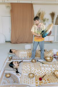 a couple lying on the floor under blankets, surrounded by singing bowls on or near them, and a person holding a gong with a mallet